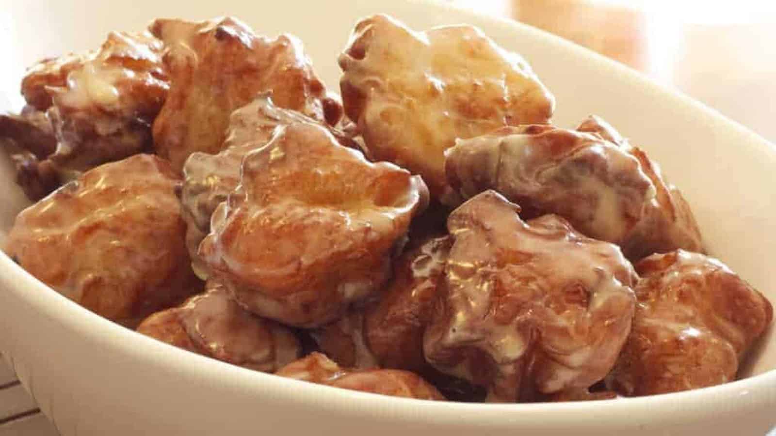 A white bowl filled with glazed apple fritters, showing their golden-brown, irregular shapes and shiny coating.