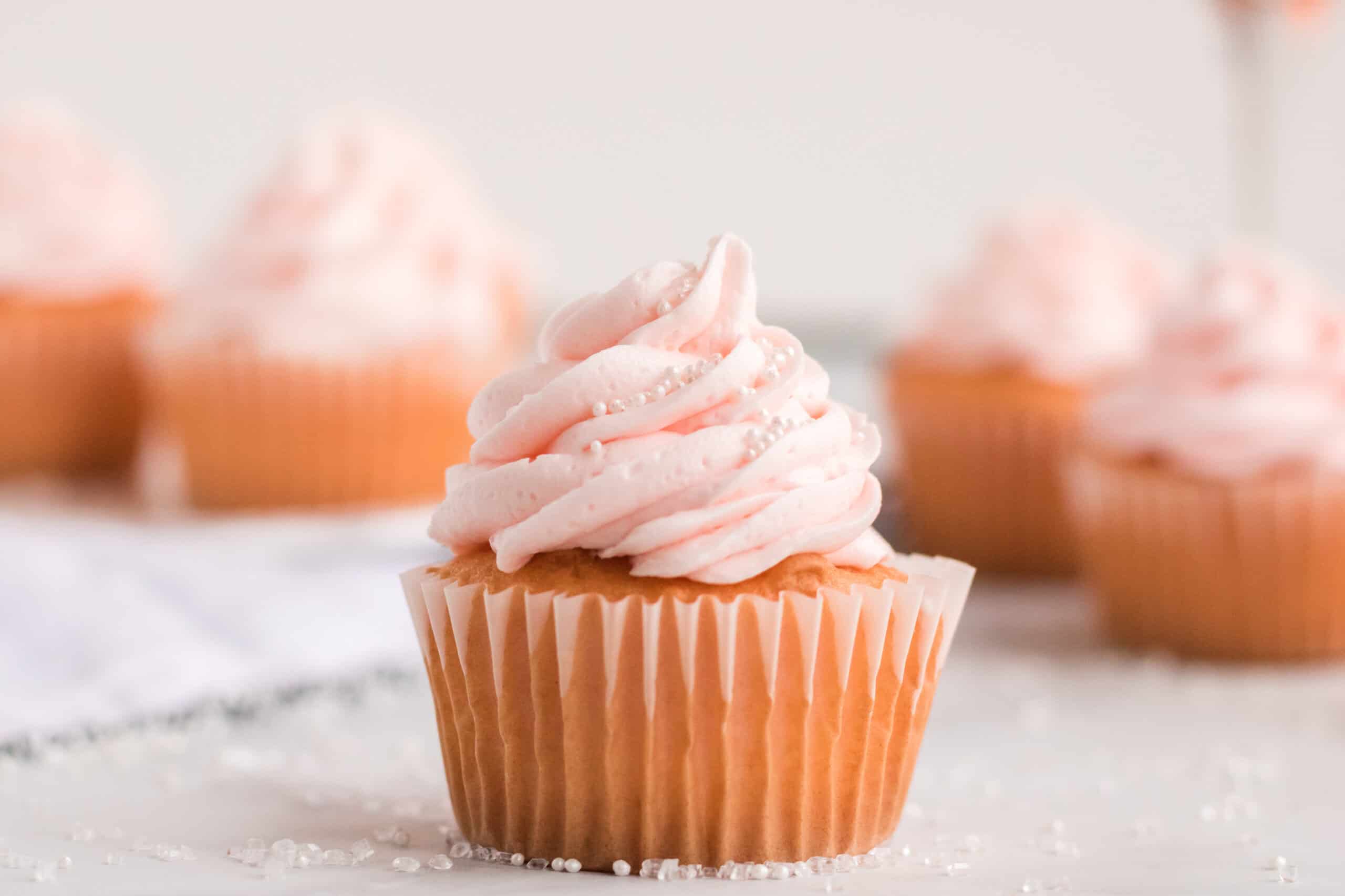 A cupcake with light pink frosting and sugar sprinkles on a white surface, with more cupcakes blurred in the background.
