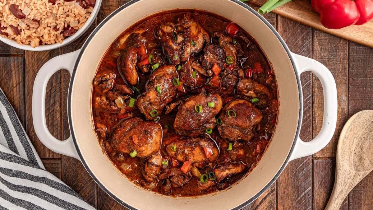 A pot of stewed chicken with vegetables and sauce, garnished with chopped green onions, sits on a wooden table next to a bowl of rice and beans.
