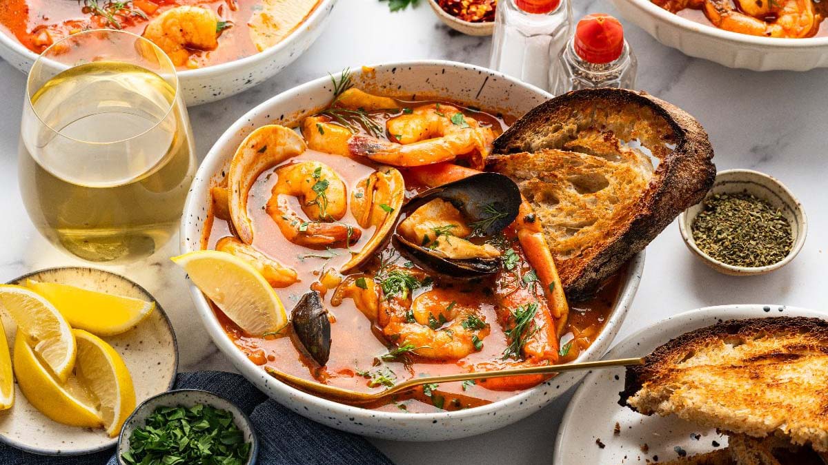 A bowl of seafood stew with shrimp, mussels, and clams in tomato broth, garnished with herbs, served with toasted bread, lemon wedges, and a glass of white wine.