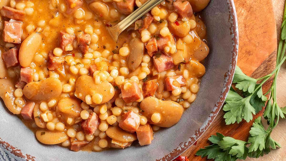 A close-up of a bowl filled with bean and sausage soup, featuring white beans, sausage pieces, small pasta, and broth, with a spoon resting in the bowl. Parsley is nearby.