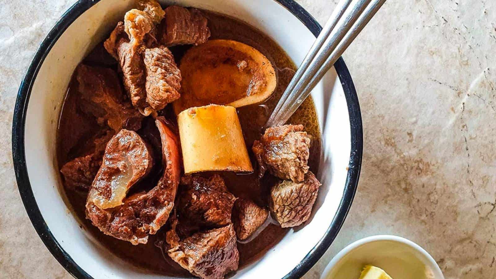 A bowl of beef stew with bone marrow and chunks of meat, served with a spoon and fork on a marble surface. A small dish with a lemon wedge is visible beside the bowl.