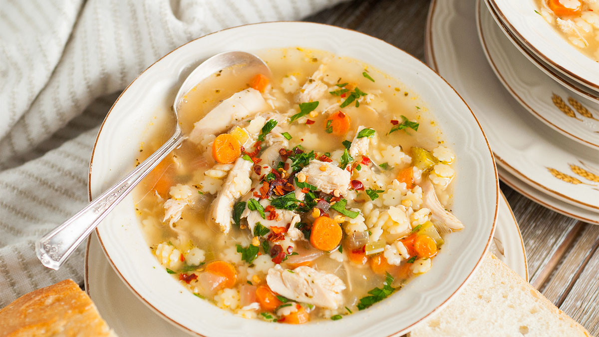 A bowl of chicken and rice soup with carrots, celery, and herbs, served with a spoon and slices of bread on the side.