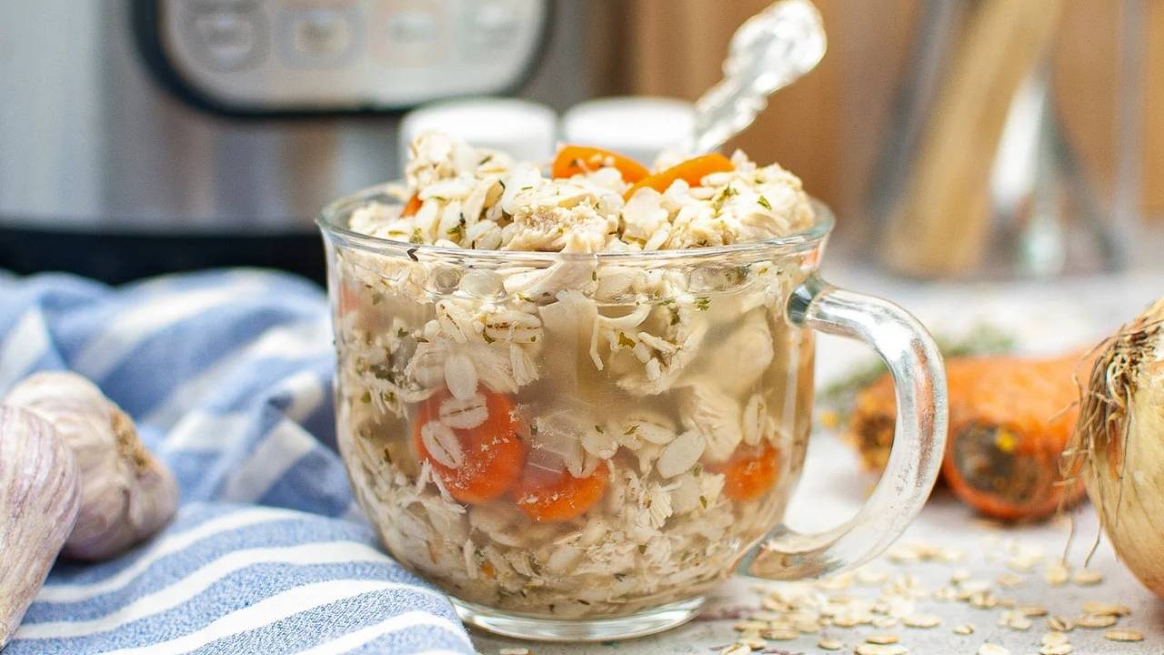 A clear glass mug filled with chicken soup containing shredded chicken, oats, carrots, and herbs, placed on a countertop with kitchen items in the background.