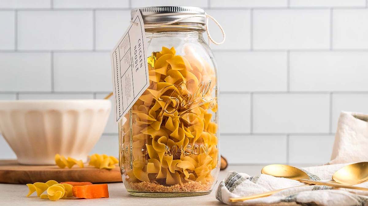 A glass mason jar filled with uncooked pasta sits on a countertop, accompanied by a recipe tag, a bowl, utensils, and a cloth.