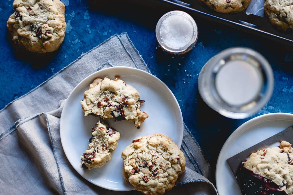A plate with chocolate chip cookies, one broken in half, sits on a napkin next to a glass of water, a jar of salt, and a baking tray with more cookies on a blue surface.