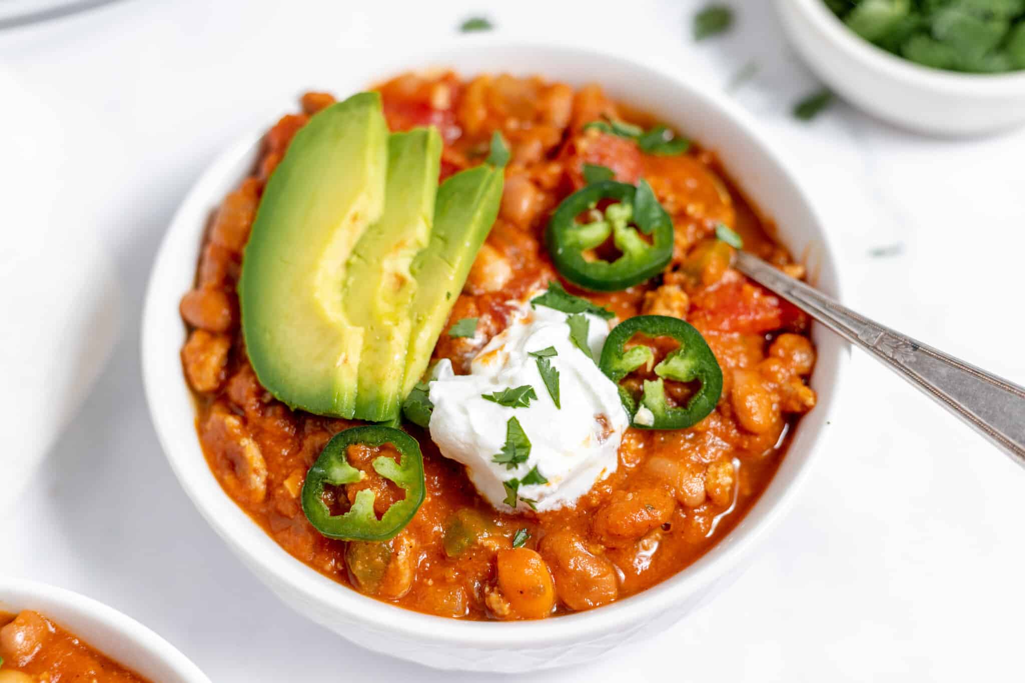 A bowl of chili topped with sliced avocado, jalapeรฑo, sour cream, and chopped cilantro, with a spoon on the side.