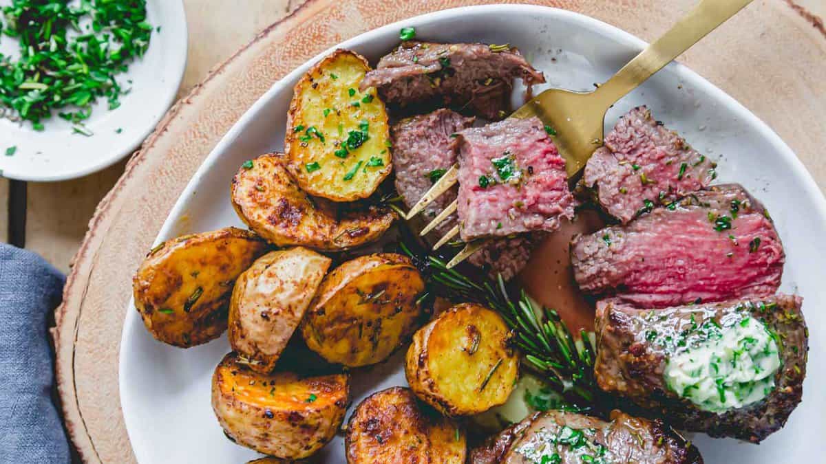 A plate of sliced medium-rare steak with a dollop of herb butter, roasted potatoes, and a sprig of rosemary, with a fork holding a piece of steak.