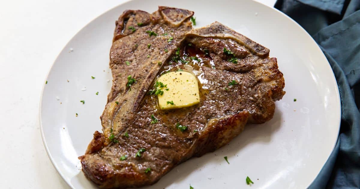 A cooked T-bone steak on a white plate, topped with a pat of melting butter and sprinkled with chopped herbs.