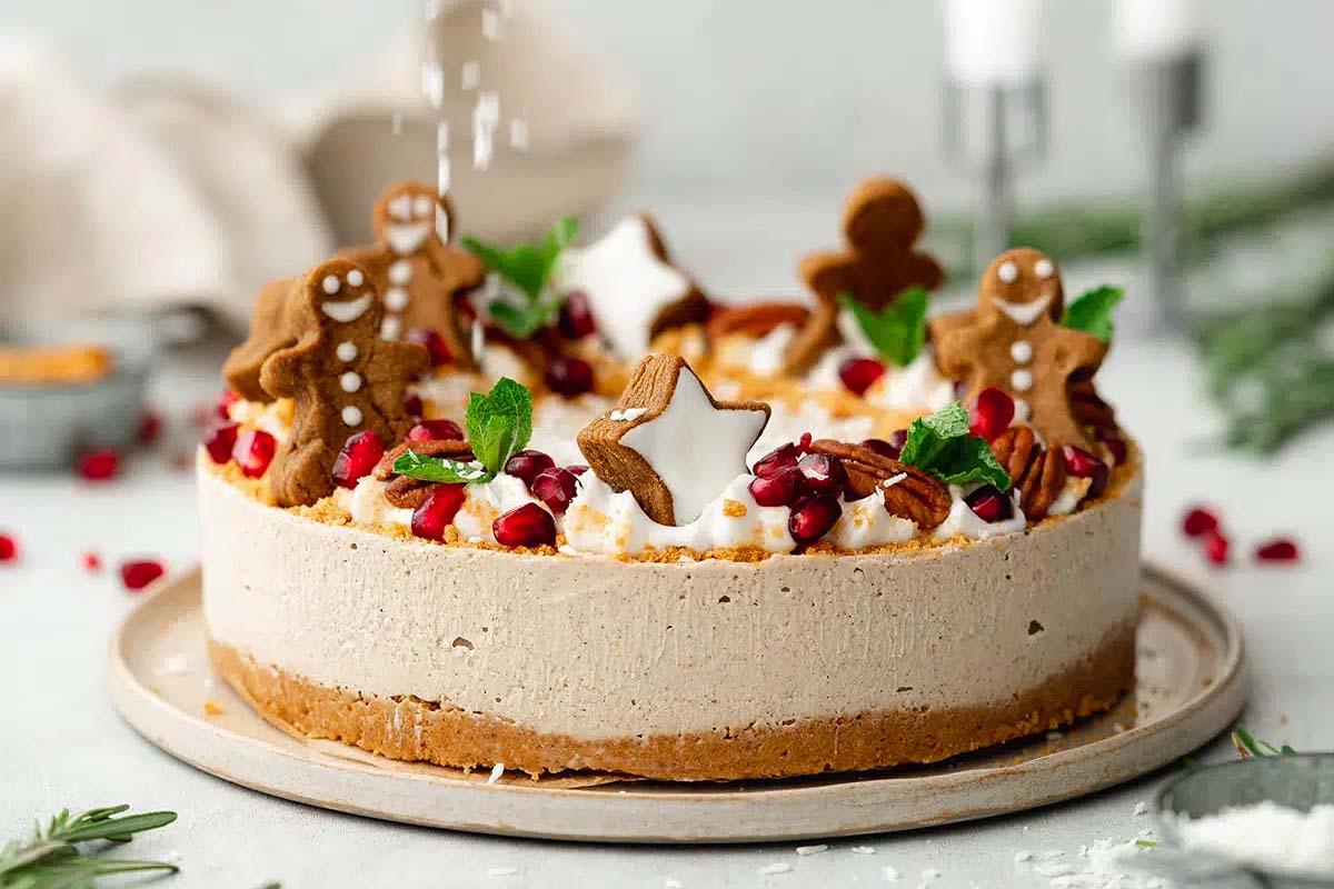 A round cake topped with whipped cream, gingerbread cookies, star-shaped cookies, pomegranate seeds, mint leaves, and chopped nuts on a light plate.