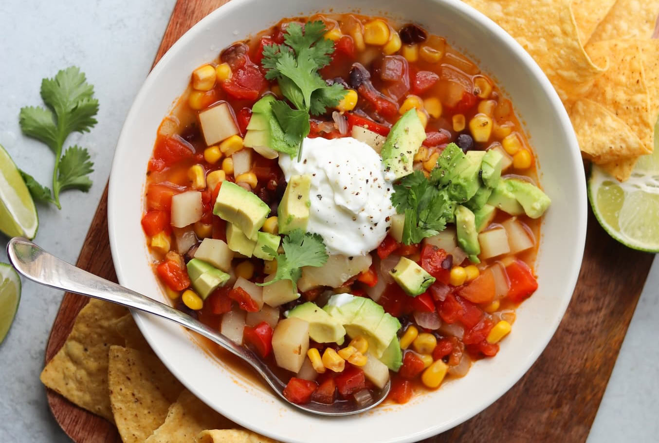 A bowl of vegetable soup with corn, tomatoes, black beans, potato, and avocado, topped with sour cream and cilantro, served with tortilla chips and lime wedges.