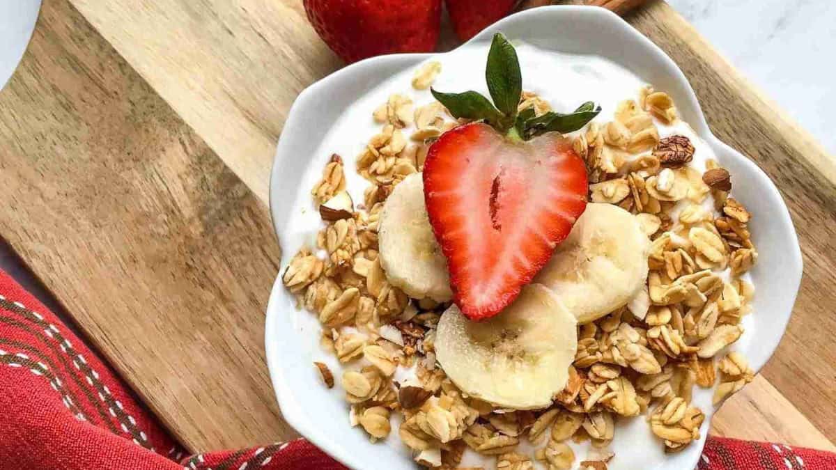 A bowl of yogurt topped with granola, banana slices, and a halved strawberry sits on a wooden board next to whole strawberries and a red cloth.