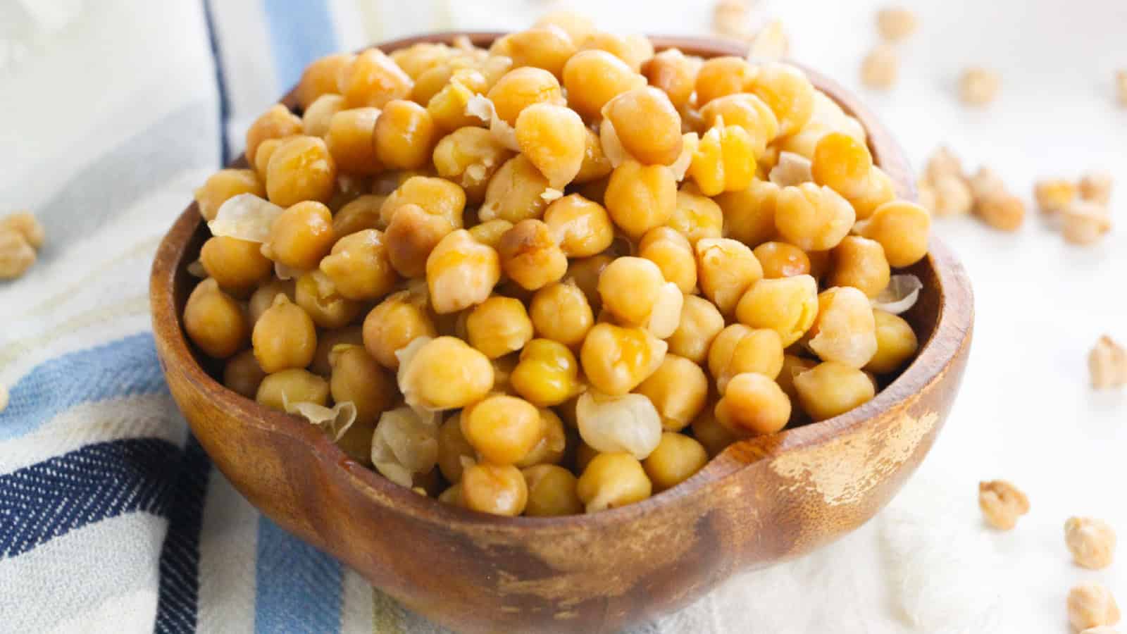 A wooden bowl filled with cooked chickpeas sits on a striped cloth, with a few loose chickpeas scattered nearby.