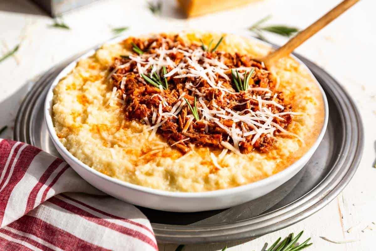 A bowl of creamy polenta topped with shredded cheese, a tomato-based meat sauce, and garnished with rosemary, placed on a silver tray with a striped red and white napkin nearby.