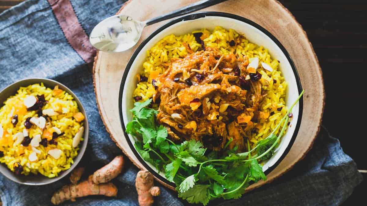 A bowl of yellow rice topped with shredded meat, dried fruits, sliced almonds, and fresh cilantro; a smaller bowl of rice and a spoon are beside it.