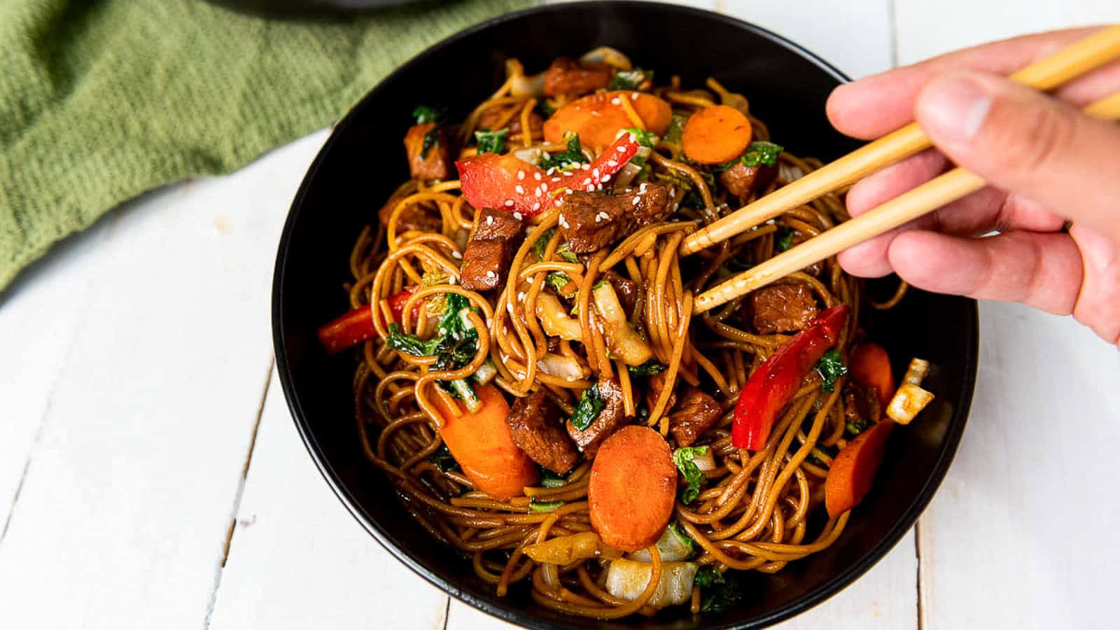 A hand uses chopsticks to pick up noodles, vegetables, and beef from a black bowl of stir-fried lo mein on a white table.