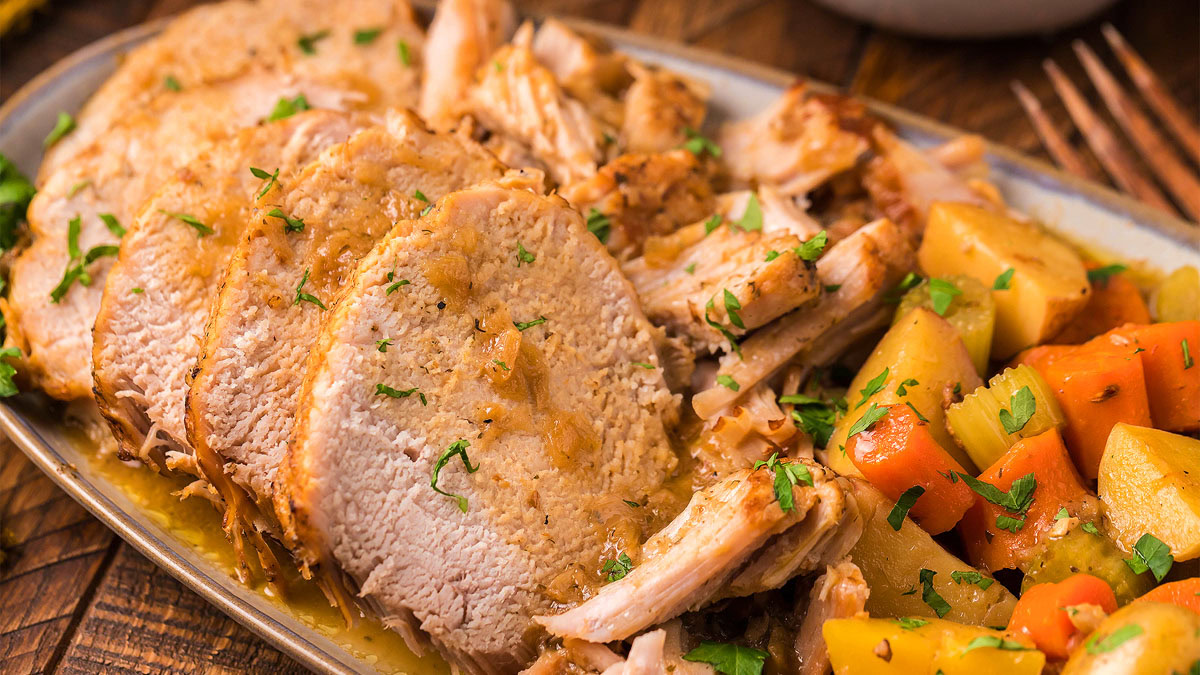 Sliced roast pork with gravy is served alongside cooked potatoes and carrots on a plate, garnished with chopped herbs.