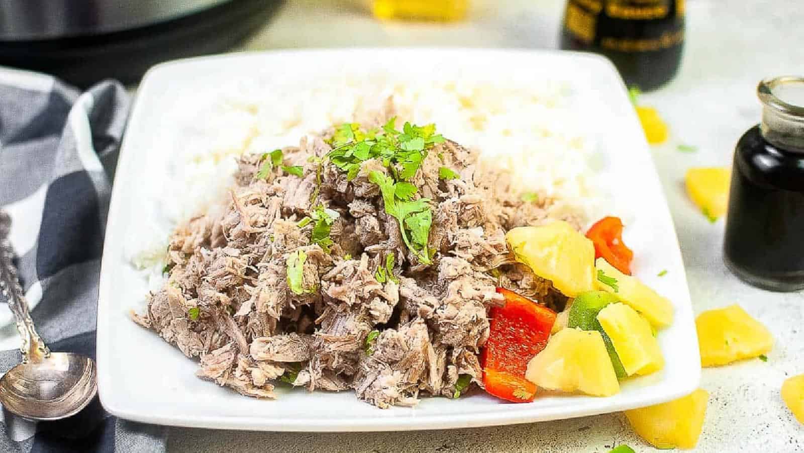 A white plate with shredded meat topped with herbs, served with white rice and a side of pineapple chunks and red bell pepper.