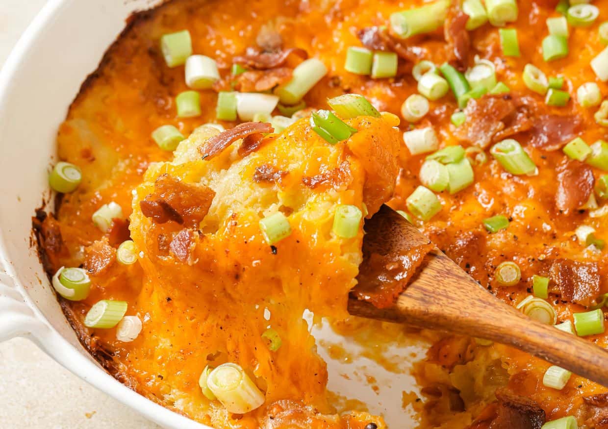 A wooden spoon scoops cheesy baked casserole topped with chopped green onions and bacon from a white dish.