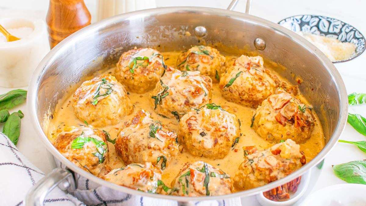A stainless steel pan filled with large meatballs in a creamy sauce, garnished with chopped herbs, sits on a white countertop.