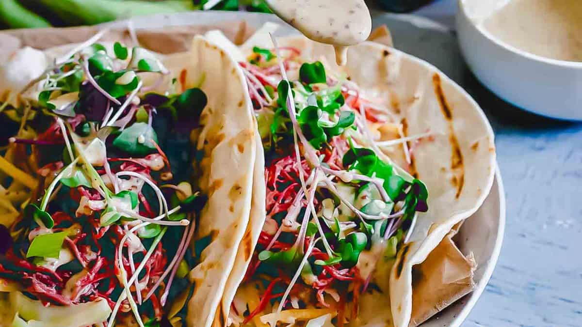Two soft tacos filled with shredded vegetables, microgreens, and cheese are drizzled with a creamy sauce, with a bowl of extra sauce visible in the background.