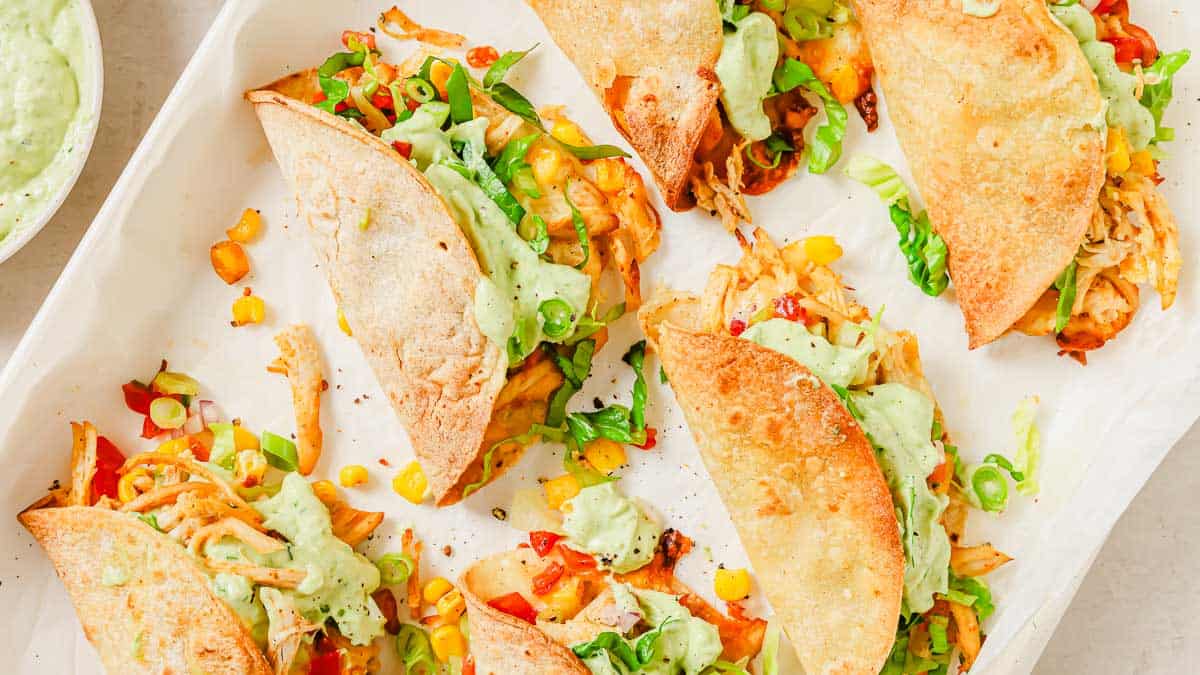 A tray of crispy tacos filled with shredded chicken, corn, diced peppers, lettuce, and topped with creamy green sauce and sliced green onions.