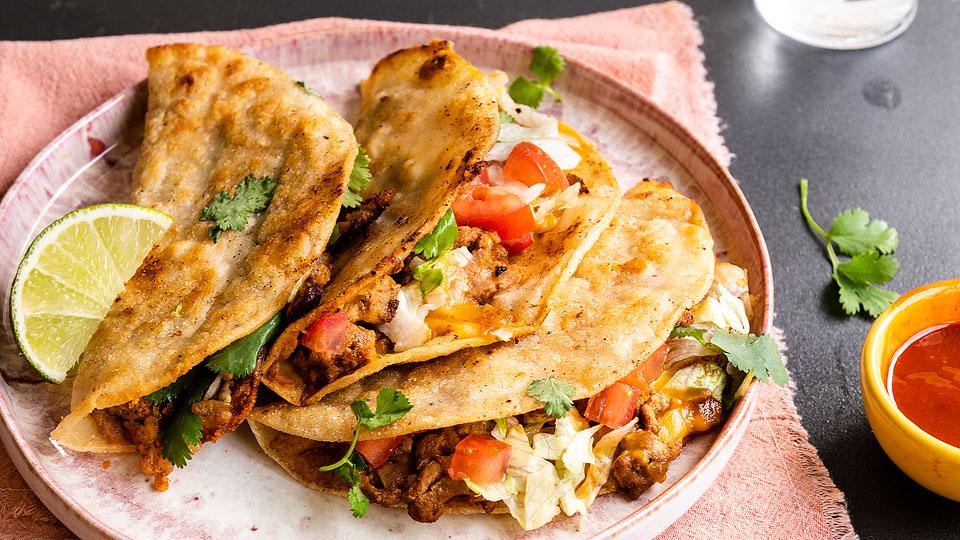 Three crispy tacos filled with meat, lettuce, tomatoes, and cilantro are served on a plate with a lime wedge, next to a small bowl of red sauce.