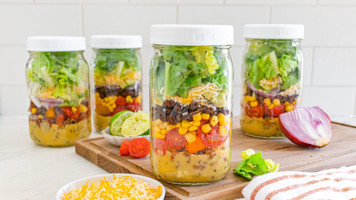 Four mason jars filled with layered salad ingredients, including lettuce, corn, black beans, tomatoes, and cheese, are arranged on a countertop with fresh produce and cheese nearby.
