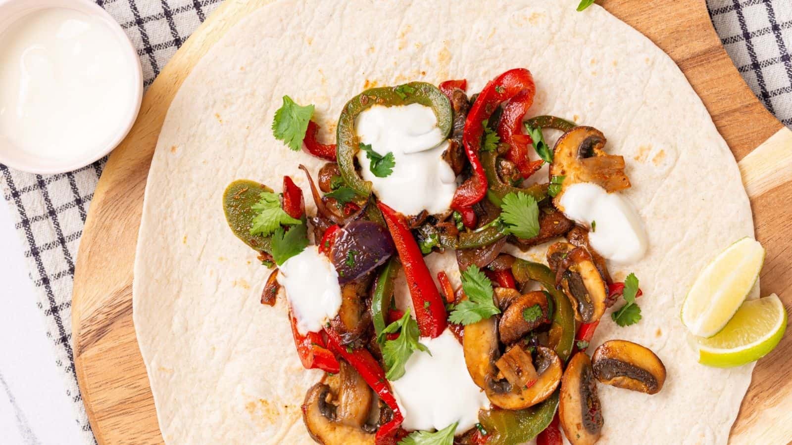 A tortilla topped with cooked mushrooms, bell peppers, onions, cilantro, and dollops of sour cream, served with lime wedges on a wooden board.