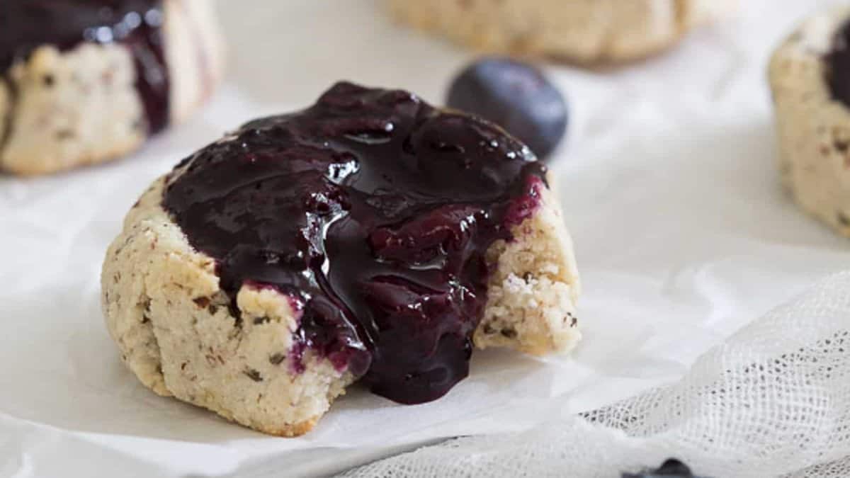 A cookie with a thick layer of dark berry jam on top, partially eaten, sits on white parchment paper.