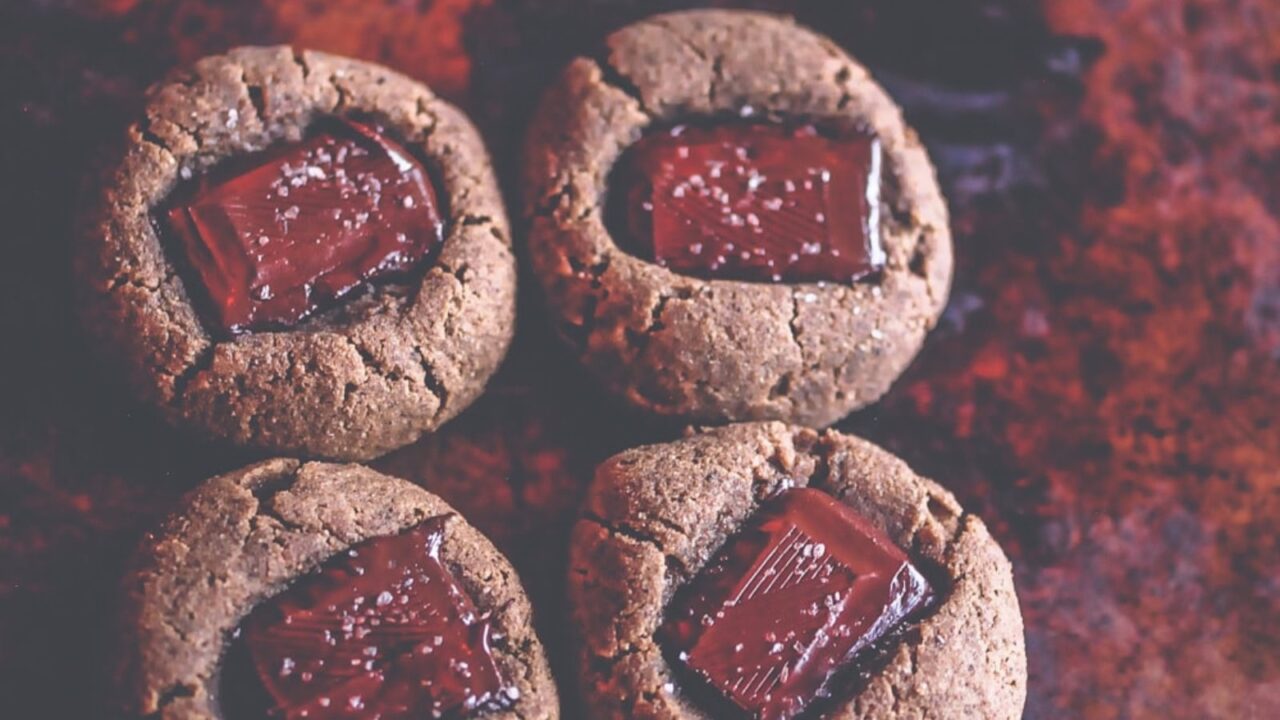 Four round cookies topped with rectangular pieces of chocolate and sprinkled with salt, arranged on a reddish-brown surface.
