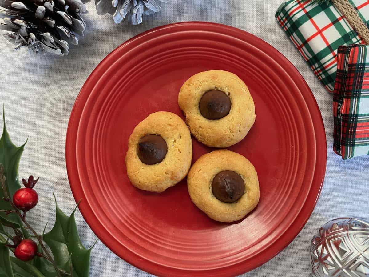 Three thumbprint cookies with chocolate centers on a red plate, surrounded by holiday decorations including holly, pinecones, and a wrapped gift.