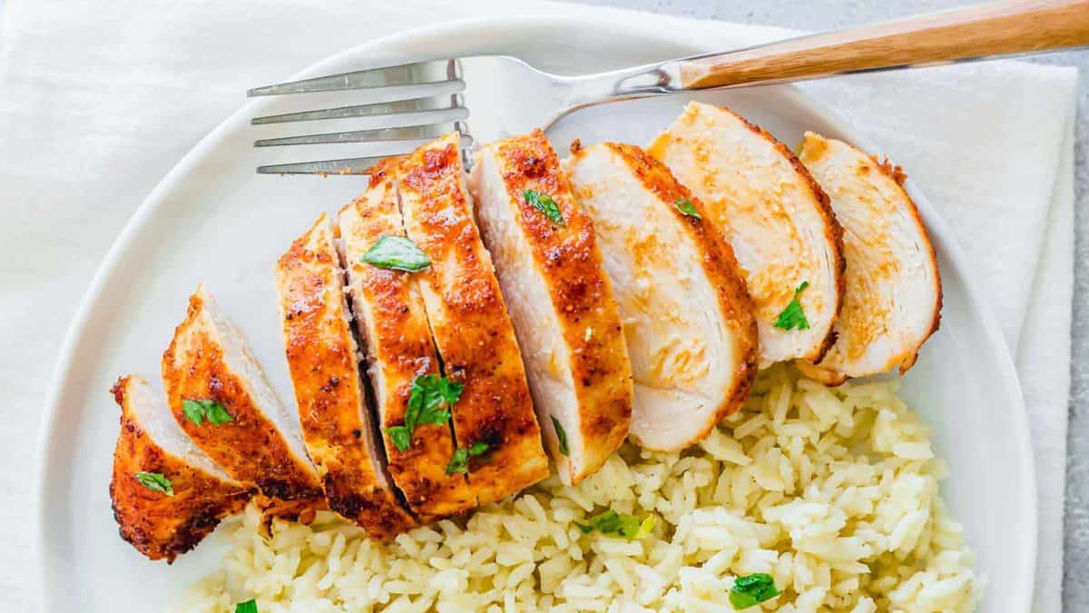 Sliced grilled chicken breast garnished with herbs served on a bed of rice, with a fork placed beside the chicken on a white plate.