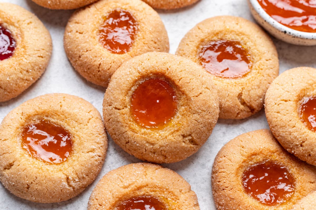 A close-up view of several round thumbprint cookies filled with orange and red jam centers, arranged on a light surface.