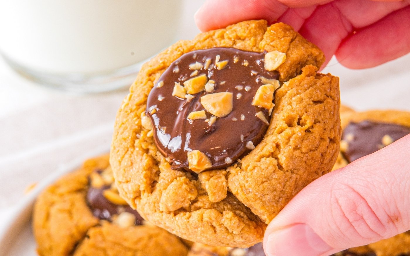 A hand holding a peanut butter cookie topped with melted chocolate and chopped peanuts; a glass of milk in the background.