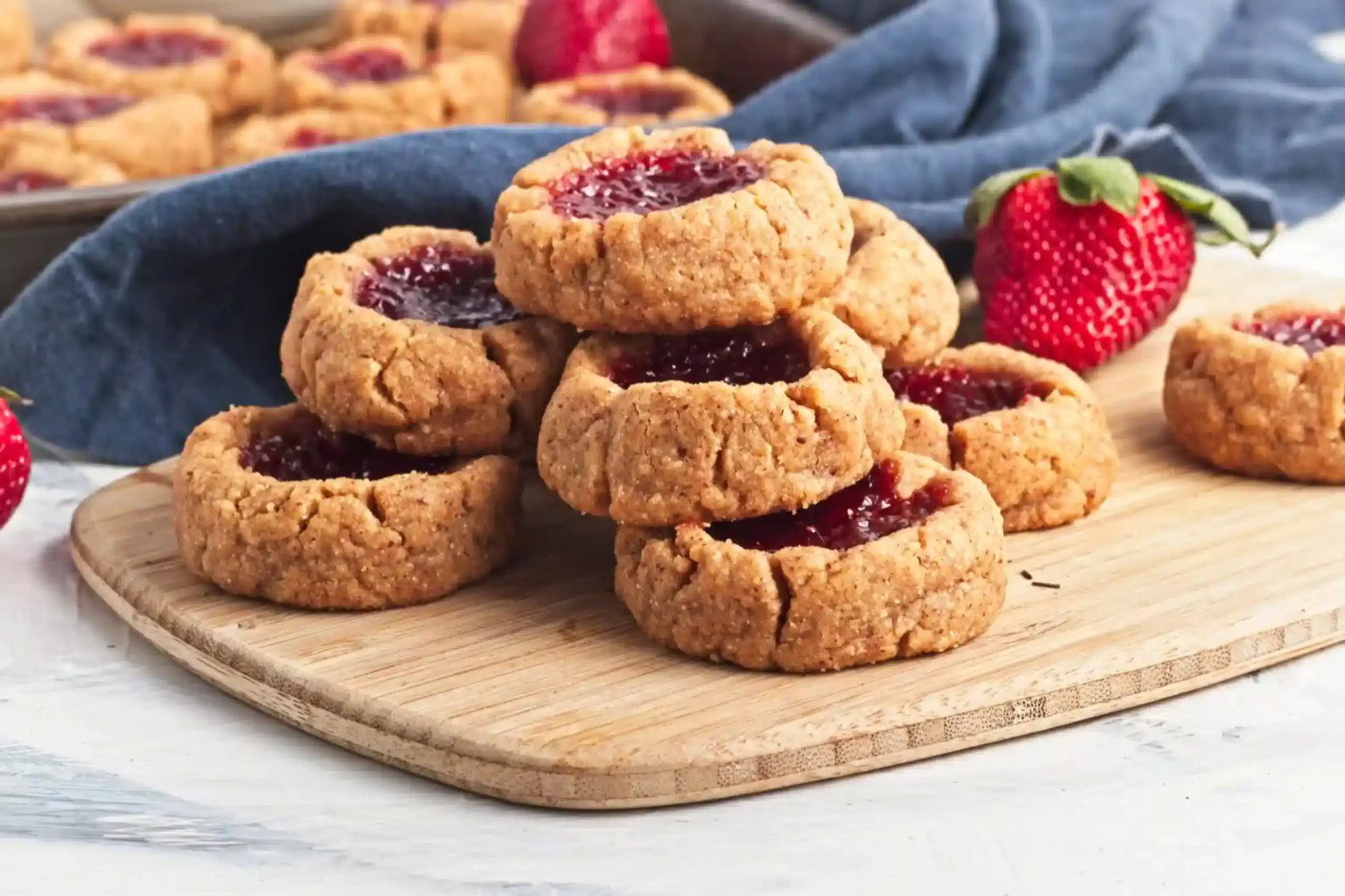 A stack of thumbprint cookies filled with red jam sits on a wooden cutting board, with fresh strawberries and more cookies in the background.