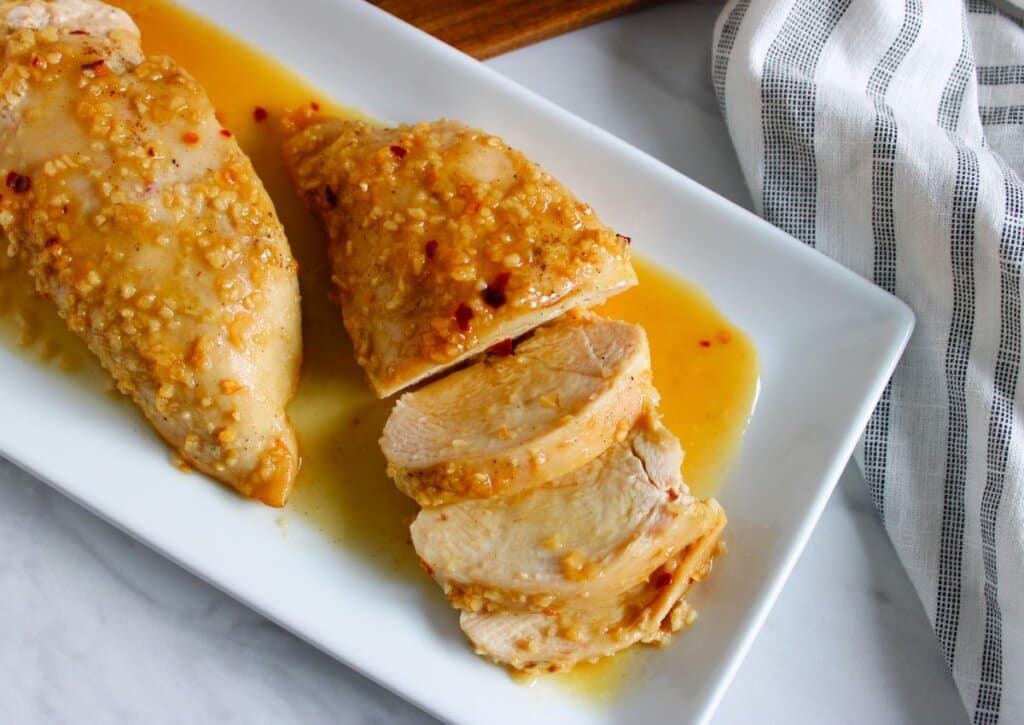 Two pieces of cooked chicken breast, one sliced, are covered in a garlic and sauce mixture on a white rectangular plate next to a striped kitchen towel.