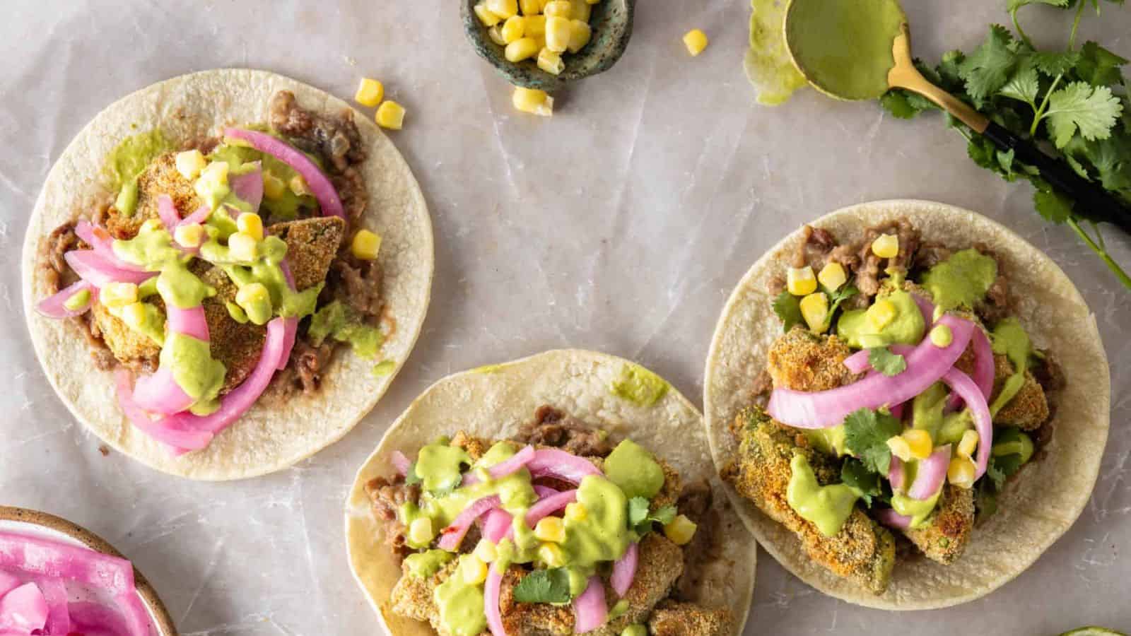 Three tacos on tortillas topped with fried vegetables, pickled onions, corn, refried beans, and green sauce, arranged on a light surface with cilantro and lime nearby.