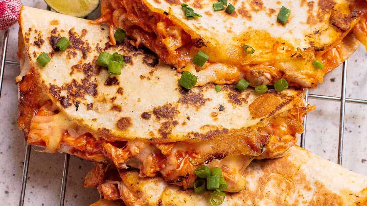 Three grilled chicken quesadillas with melted cheese and chopped green onions are stacked on a cooling rack.
