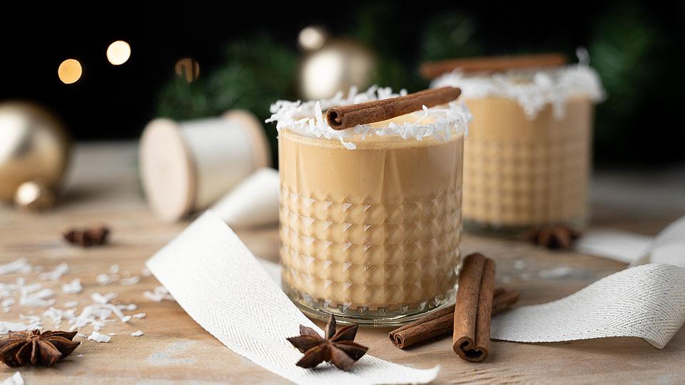 Two glasses of creamy beverage topped with shredded coconut and a cinnamon stick, surrounded by cinnamon sticks, star anise, and festive decorations on a wooden surface.