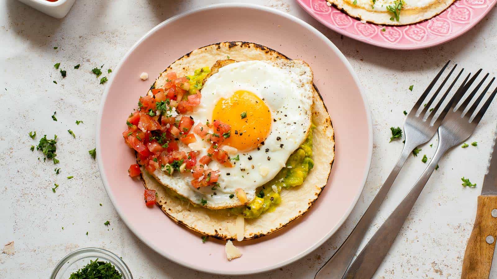 A plate with a tostada topped with guacamole, pico de gallo, and a sunny-side up egg, garnished with herbs. A fork and knife are placed beside the plate.