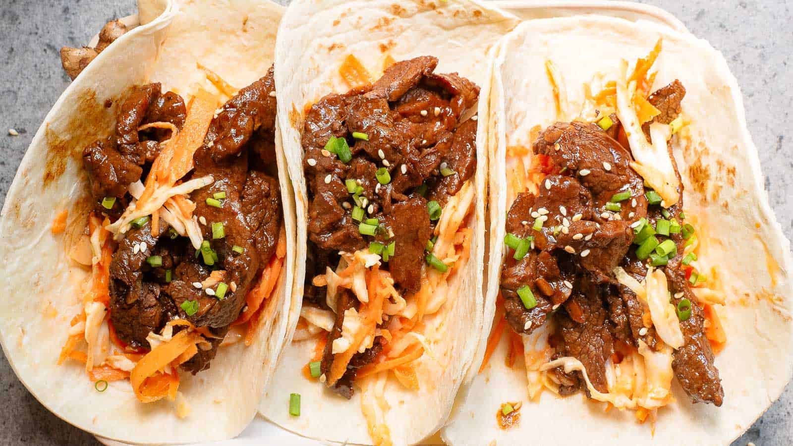 Three flour tortillas filled with marinated beef, shredded vegetables, chopped green onions, and sesame seeds, arranged side by side on a plate.