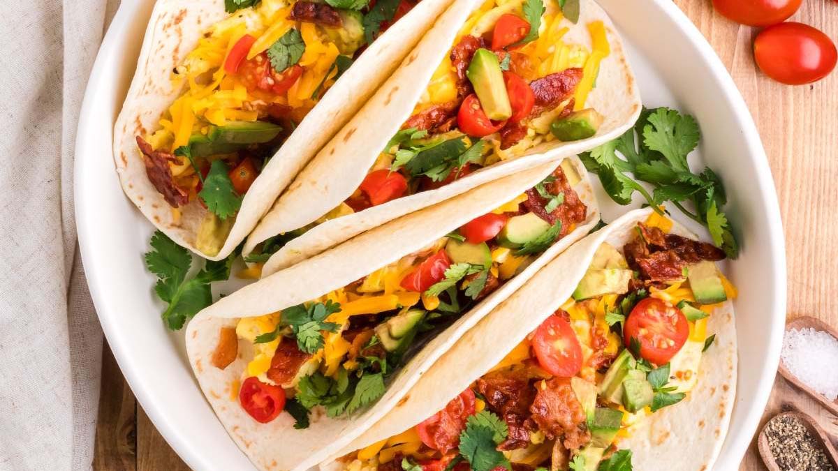Three soft tacos filled with shredded cheese, avocado, cherry tomatoes, cilantro, and other fresh ingredients, served on a white plate.