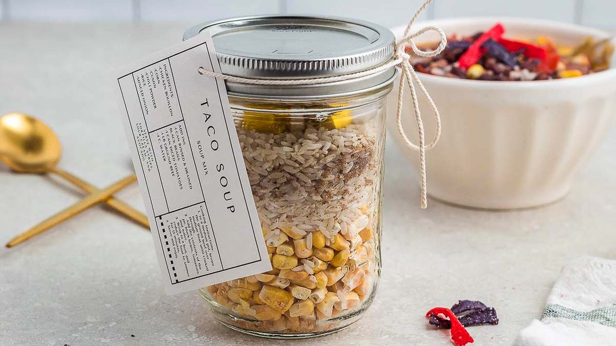 A glass jar filled with layered dry ingredients for taco soup, labeled with a recipe card, sits beside a gold spoon and a bowl of food on a countertop.