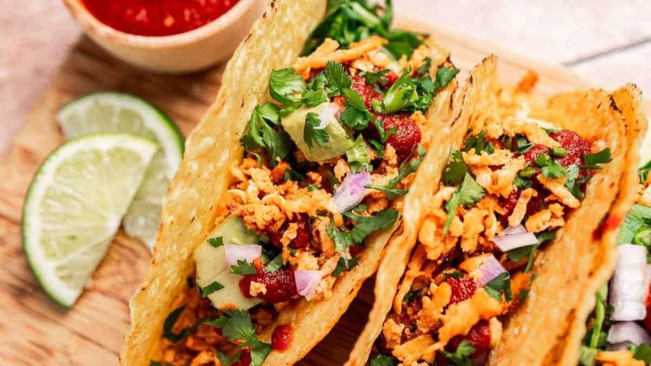 Three tacos filled with shredded vegetables, cilantro, diced onions, and sauce, served on a wooden board with lime wedges and a bowl of red salsa.