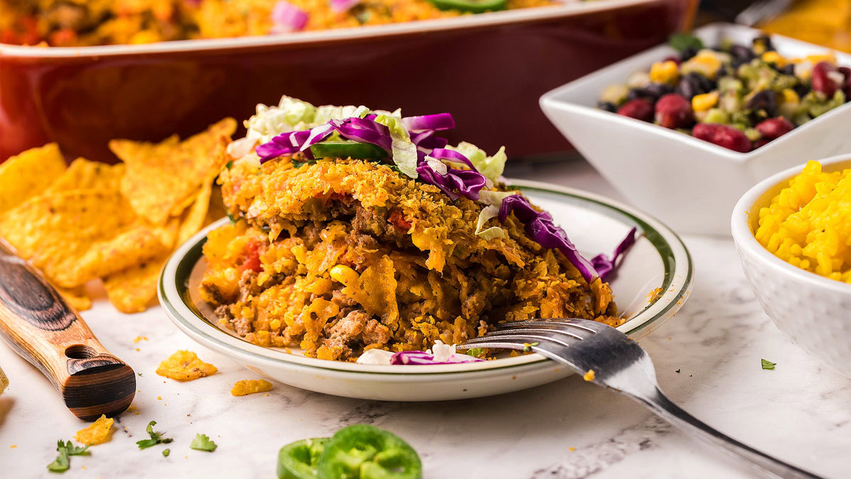 A plate of taco casserole topped with lettuce and cabbage, accompanied by tortilla chips, a fork, a bowl of yellow rice, and a bowl of bean salad.