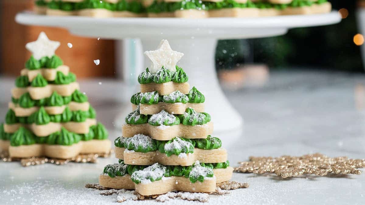 A stack of star-shaped cookies decorated with green icing and powdered sugar, arranged to resemble a Christmas tree, sits on a table with more cookie trees in the background.