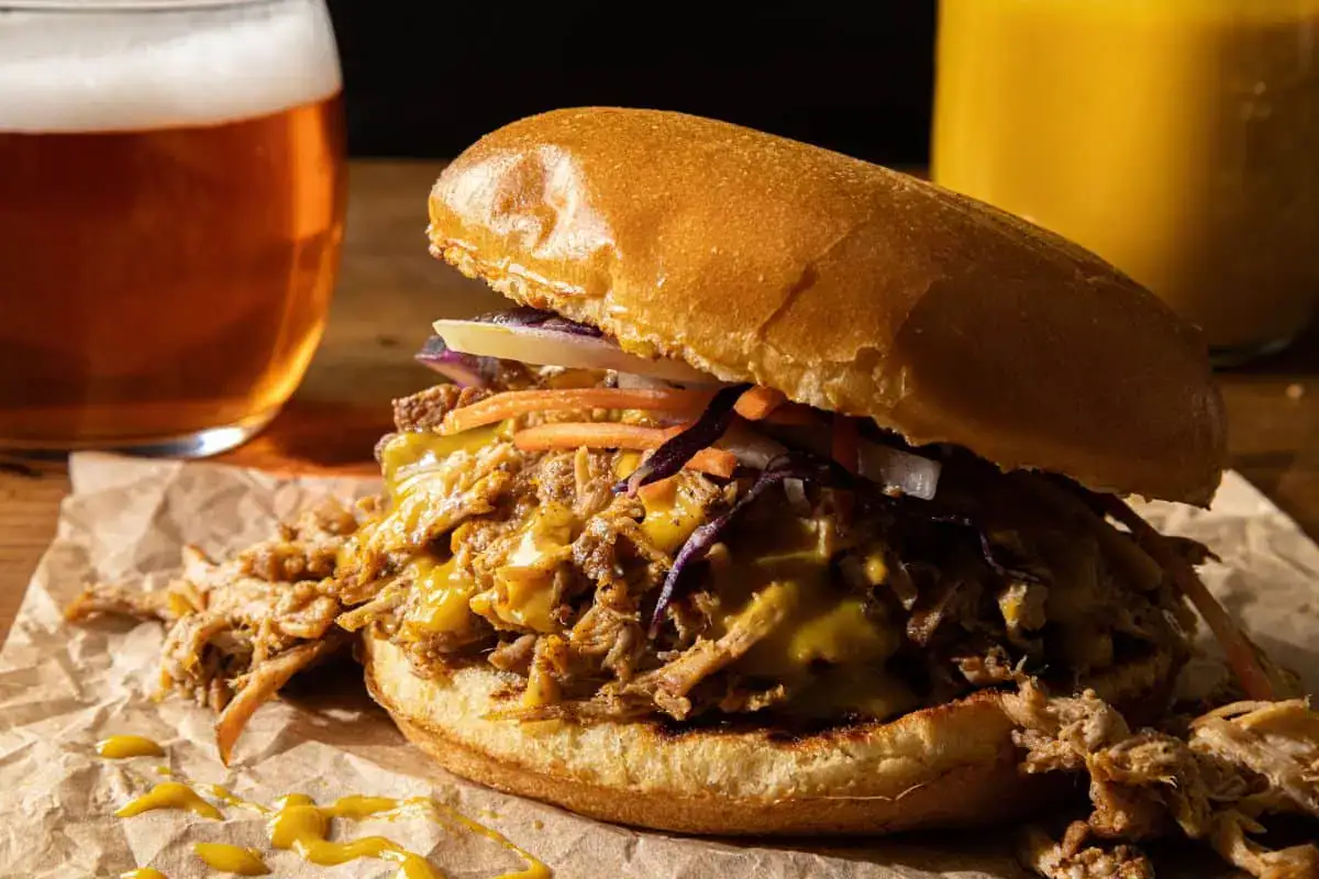 A pulled pork sandwich with melted cheese and slaw sits on parchment paper, next to a glass of beer and a yellow beverage in the background.
