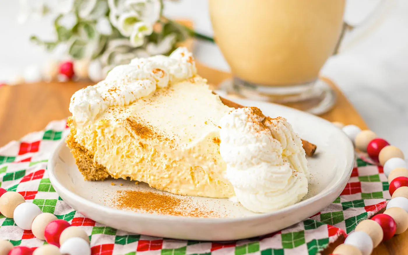 A slice of creamy pie with whipped cream and cinnamon on top sits on a white plate, with a drink and festive decorations in the background.
