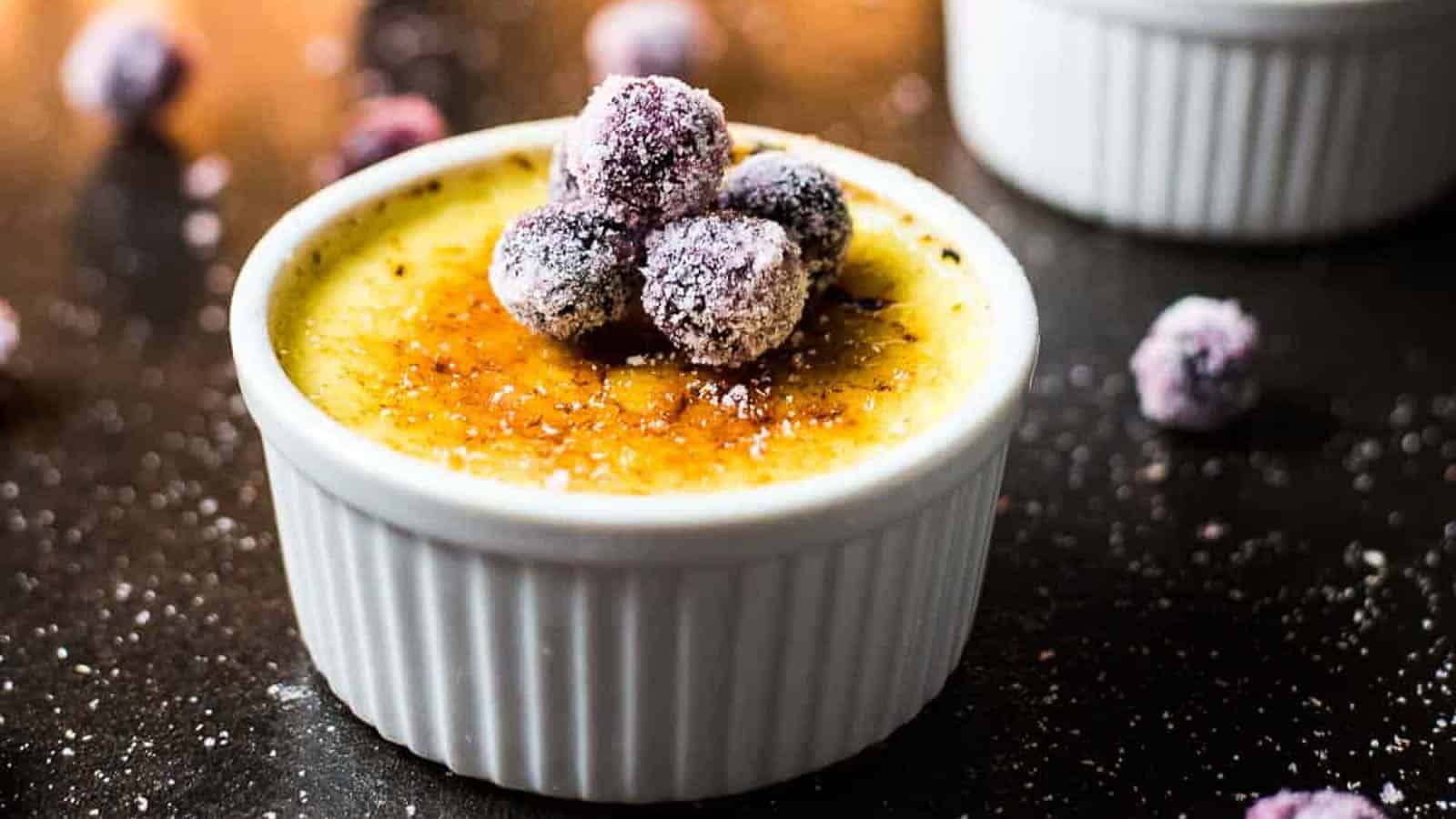 A ramekin of crรจme brรปlรฉe topped with sugared berries sits on a dark surface, with a few berries scattered around.